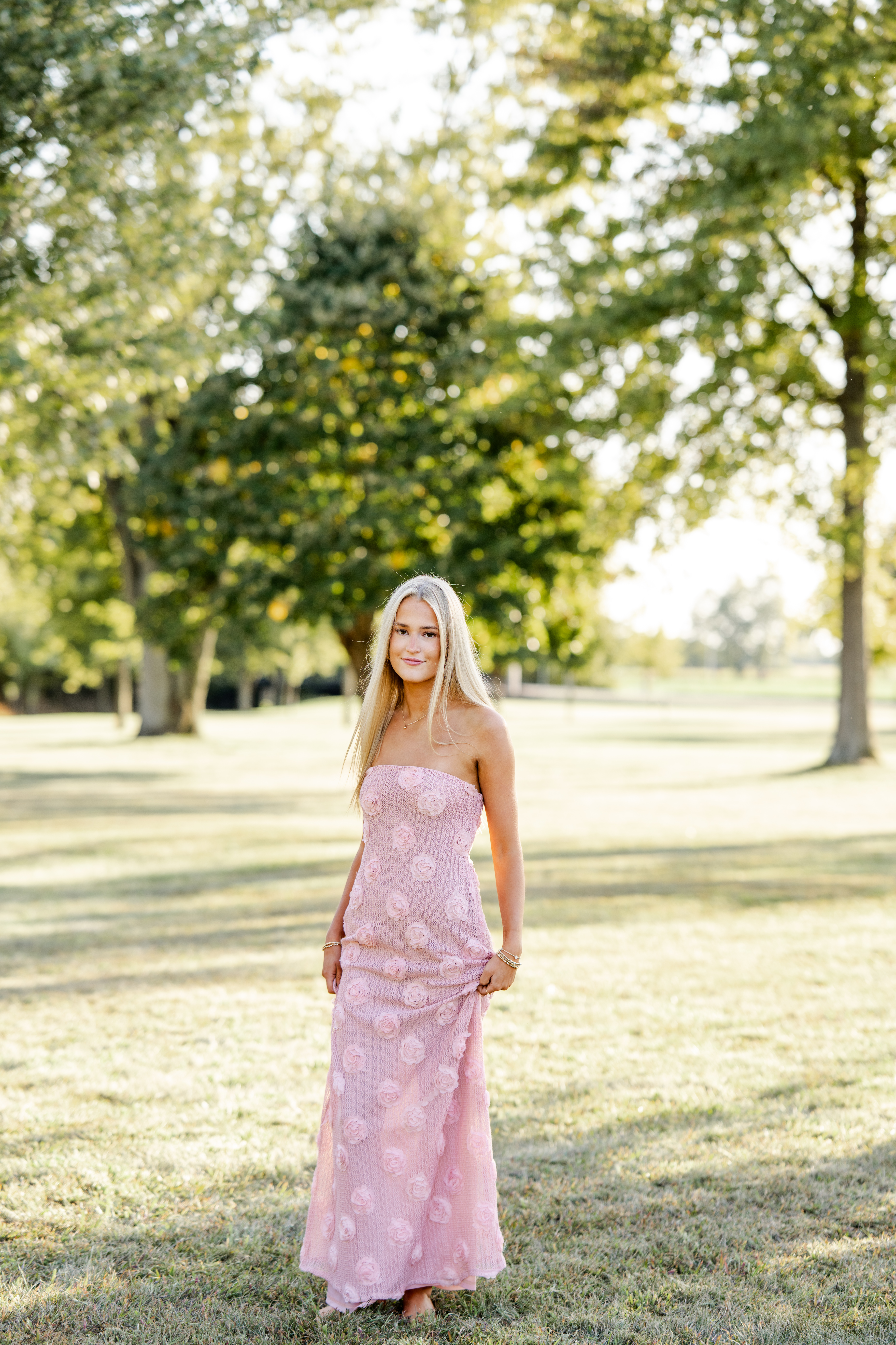 High school senior twirls in pink dress barefoot in the grass during golden hour portraits in Sheridan, Indiana.