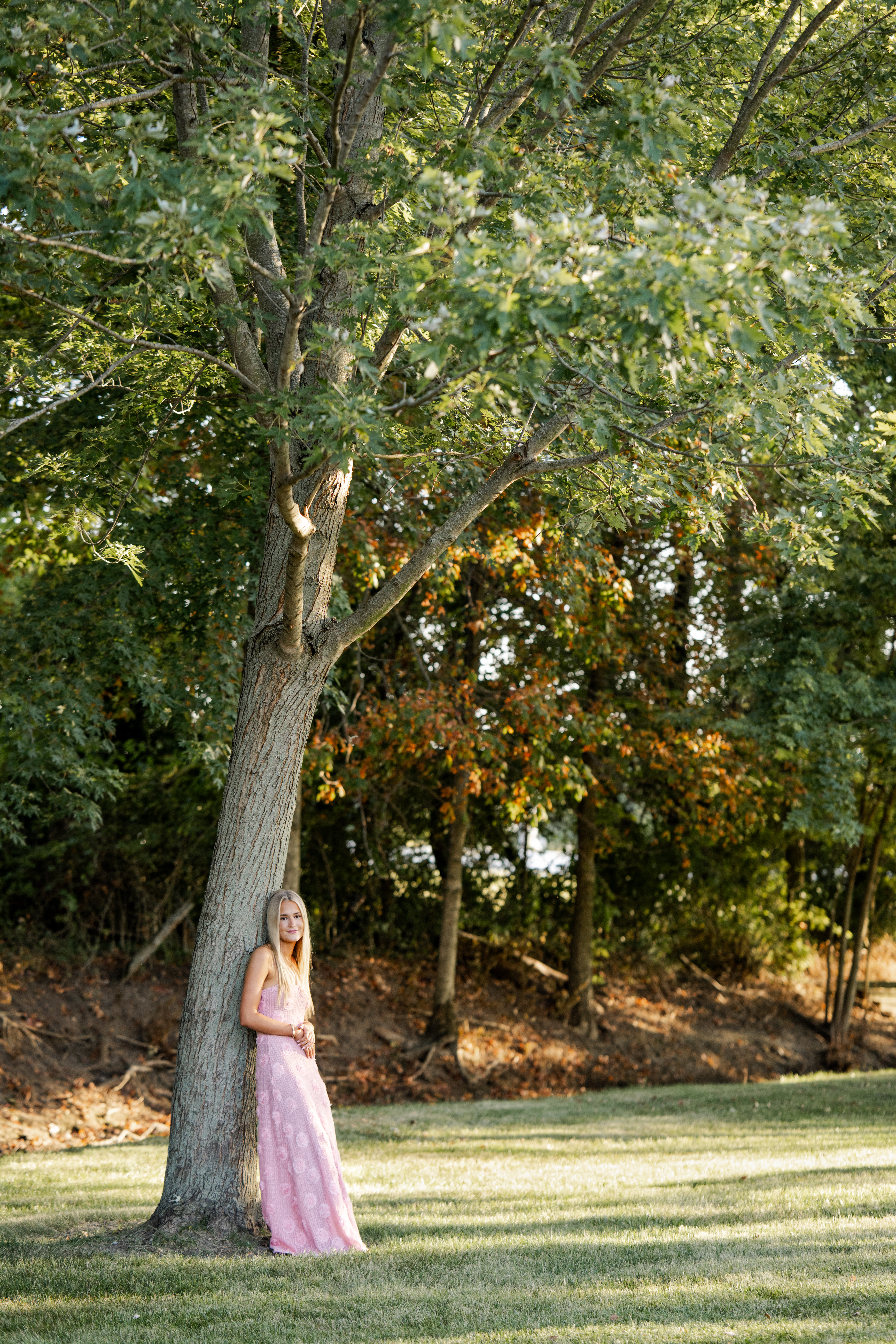 Senior girl poses beneath large tree during golden hour equestrian photos at her family farm.