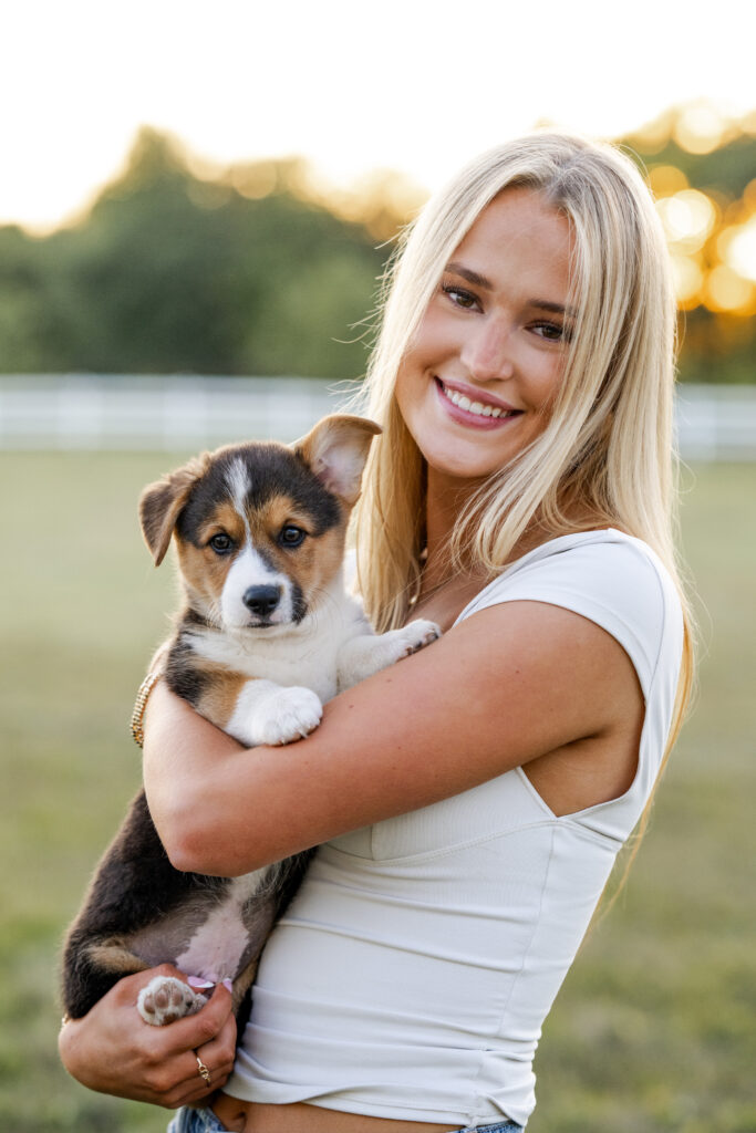 Teen girl smiles while holding corgi puppy during senior session at her family farm in Sheridan, Indiana.
